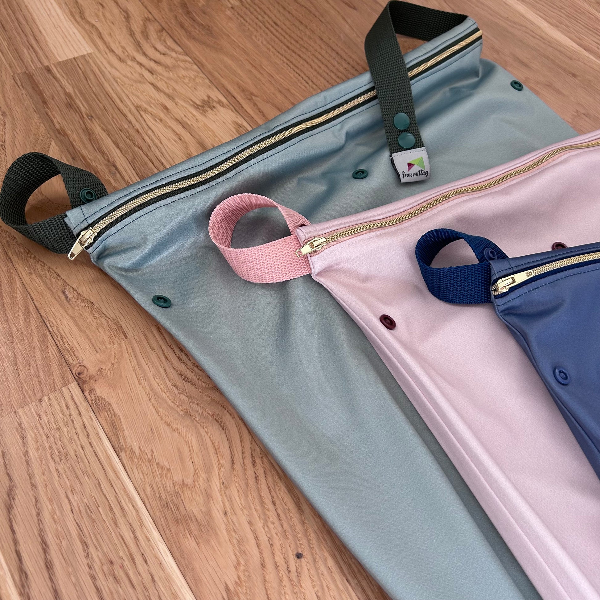 Nahaufnahme schräge Teilansicht von 3 Wetbags in salbei, nude und denim mit goldenen Reißverschlüssen. Alle drei liegen leicht versetzt auf einem Holzfussboden, frau mittag Label am oberen grünen Gurtende sichtbar.